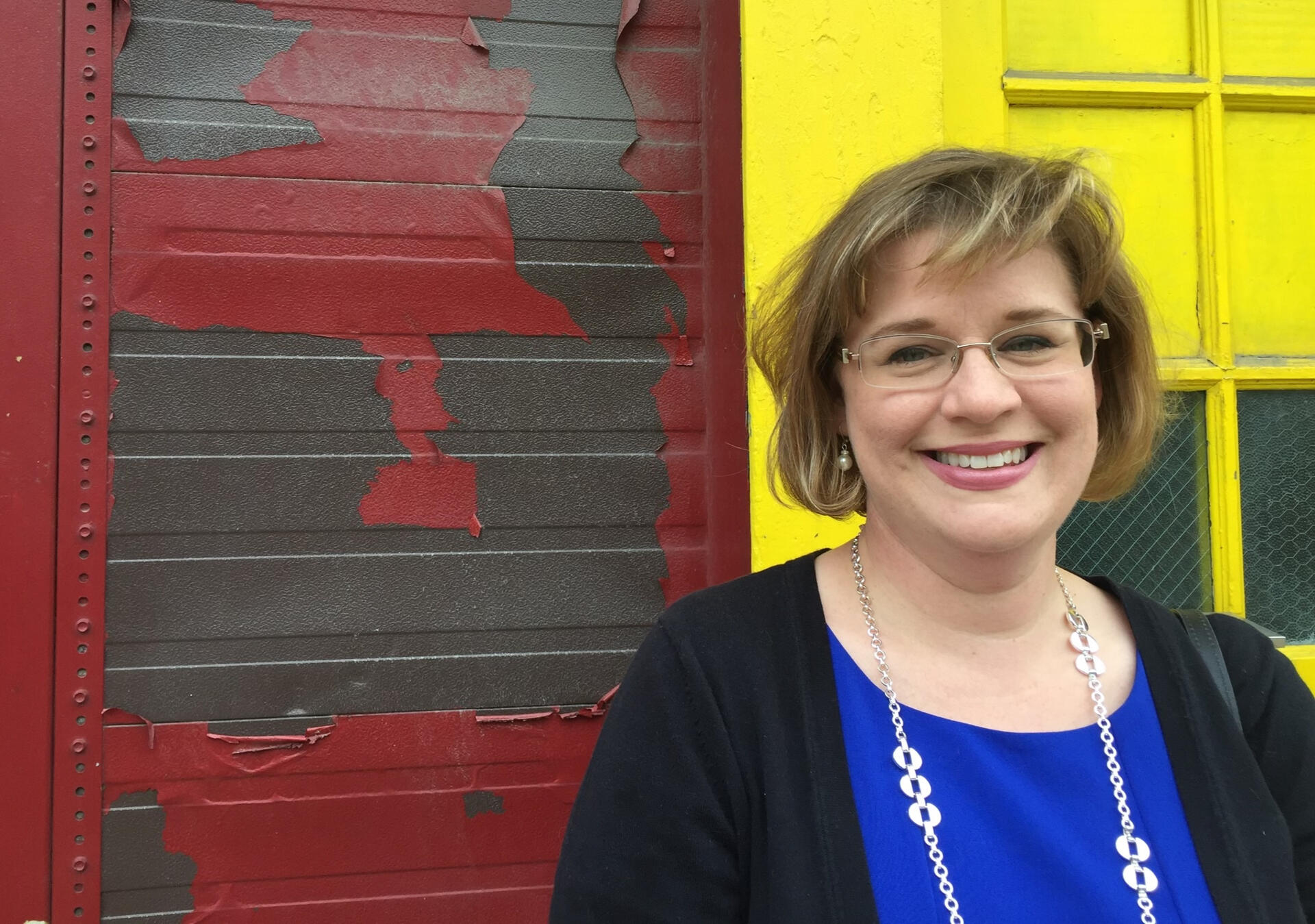 Jennifer wearing a bright blue dress and black sweater, smiling in front of an industrial looking wall in Chicago's west loop neighborhood